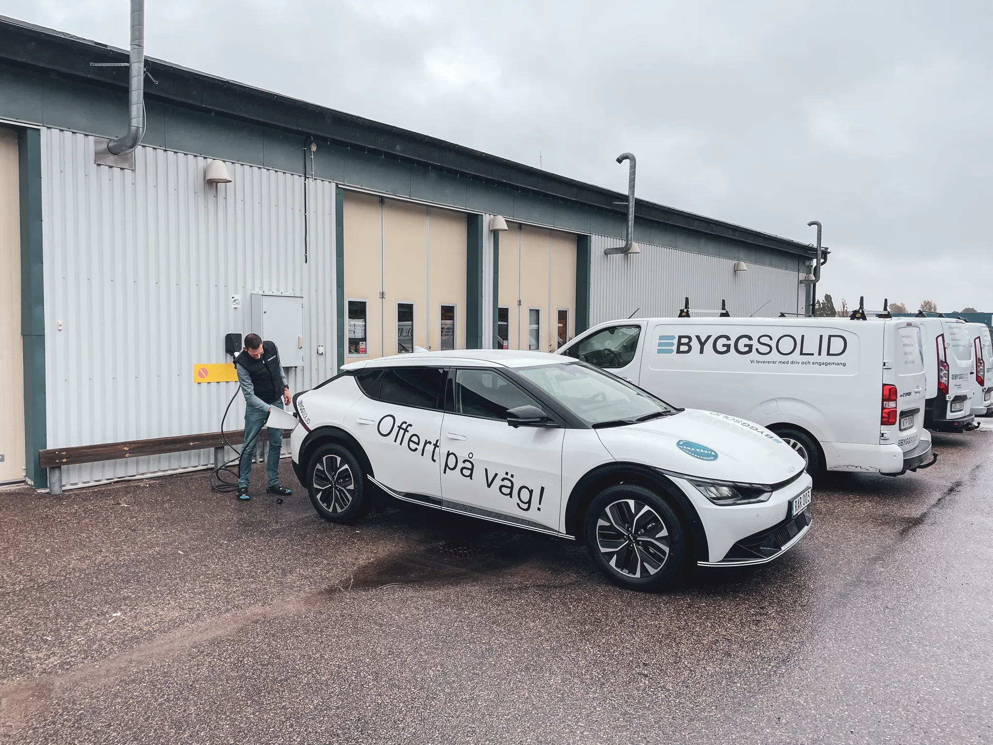 En elbil med texten Offert på väg laddas utanför Byggsolids lokaler, servicebilarna står bredvid